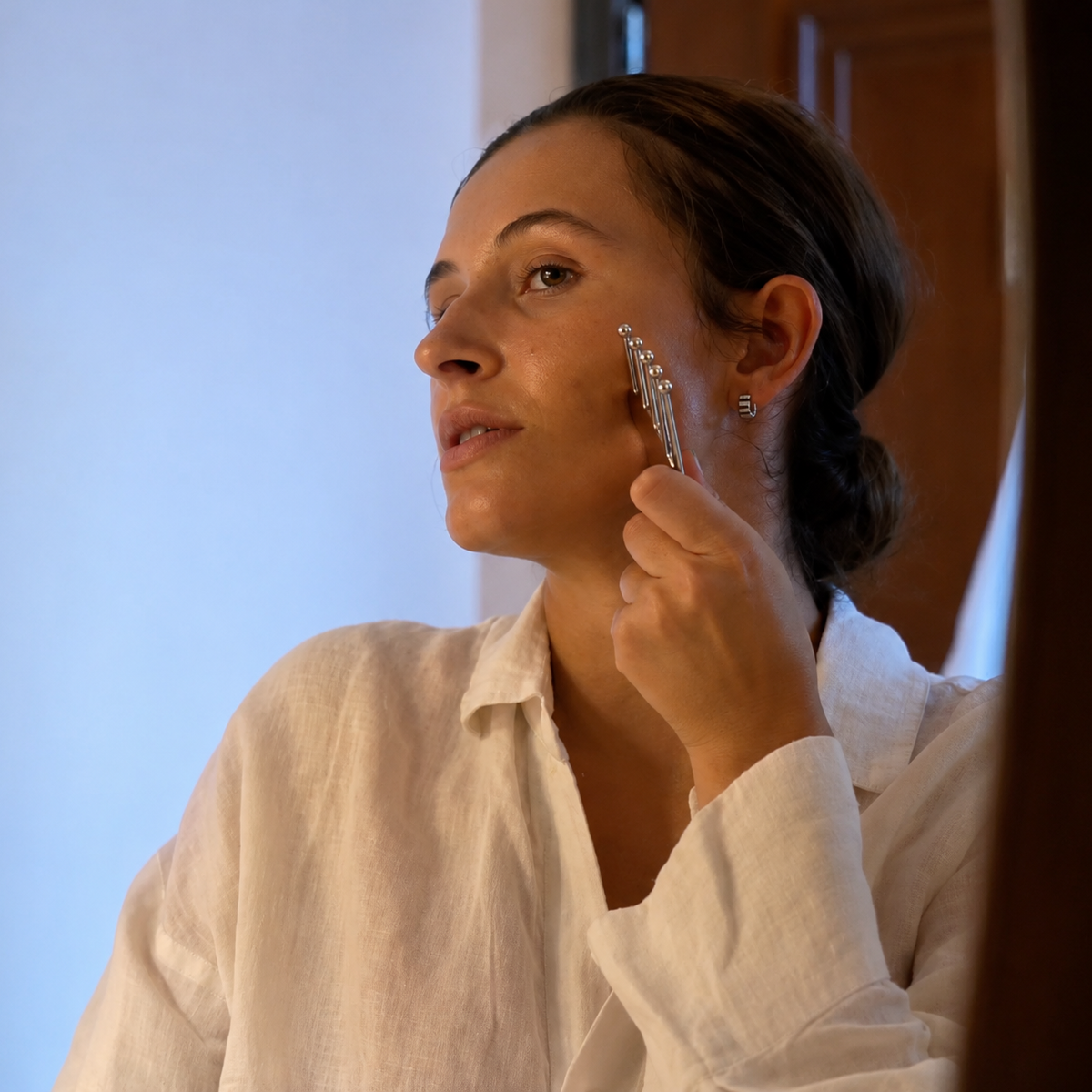 A woman in a white linen shirt uses The Daily Press Gua Sha massage tool on her cheek in a brightly lit indoor setting.
