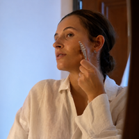 A woman in a white linen shirt uses The Daily Press Gua Sha massage tool on her cheek in a brightly lit indoor setting.