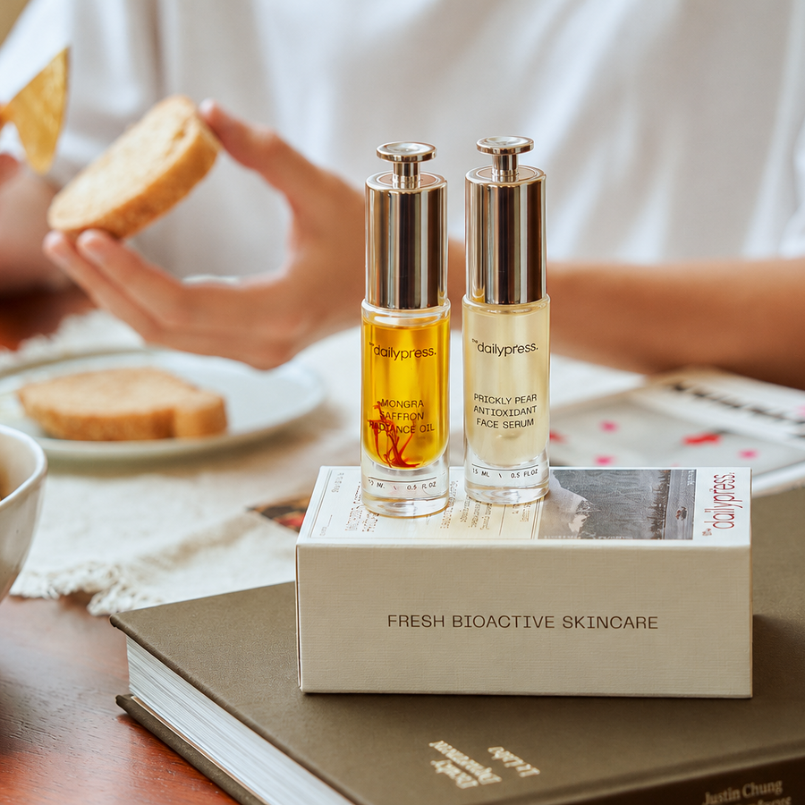 Two clear cosmetic bottles with metallic caps stand on a white box labeled 'FRESH BIOACTIVE SKINCARE' on a table. A person in a white shirt holds toast in the background, with lemons and books around them.